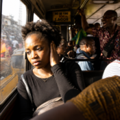 a young Black woman sitting inside a crowded Lagos bus during rush hour, staring out the window while covering one ear slightly. Bright sunlight hits her face harshly, people are talking around her, conductors shouting in the background, but her expression is emotionally distant and overwhelmed rather than dramatic.
